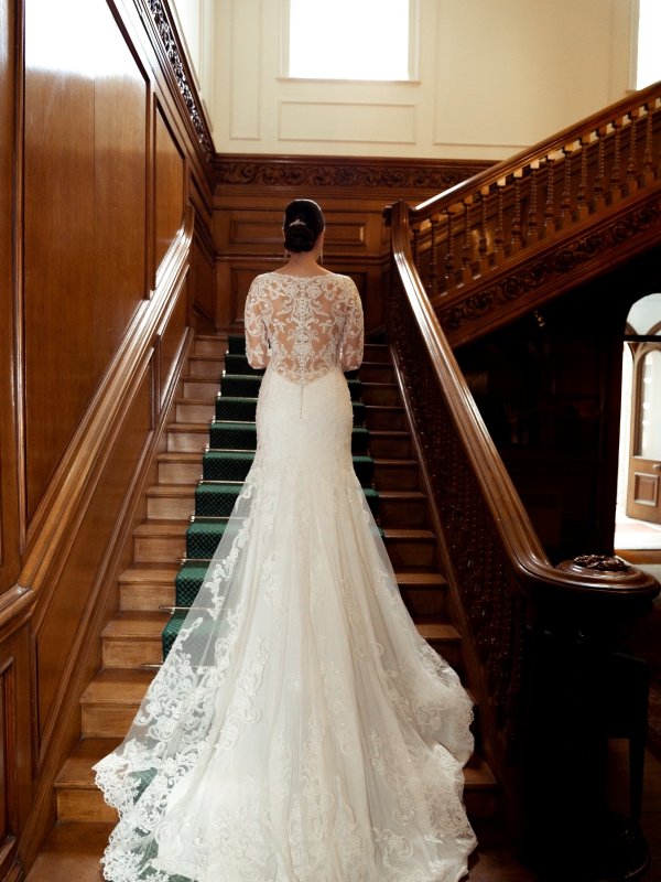 Rear View Of A Bride In A Mermaid Style Lace Gown Walking Up A Grand Wooden Staircase.